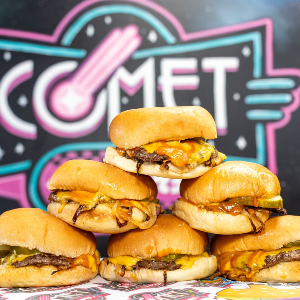 Stack of cheeseburgers with toasted buns, melted cheese, pickles, and grilled onions against a colorful neon restaurant sign.