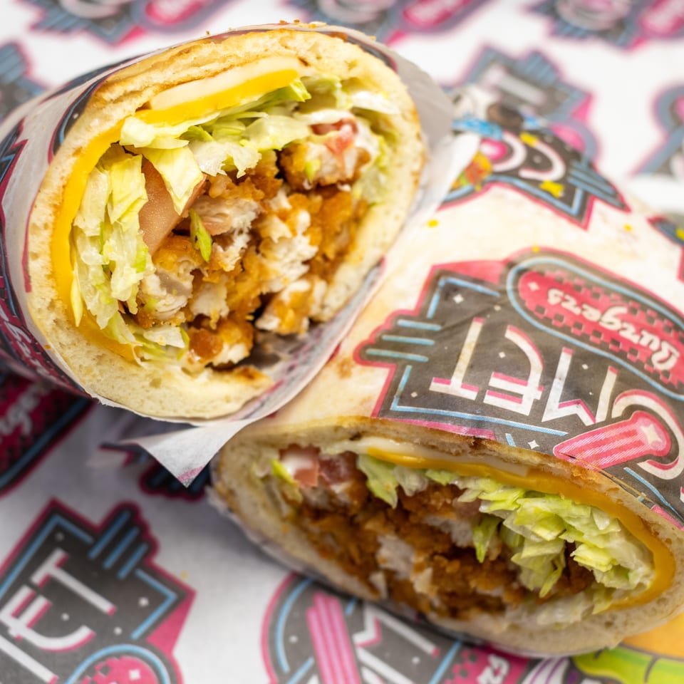 Two toasted chicken sandwiches with lettuce, tomato, cheese, and creamy sauce, wrapped in colorful branded paper.