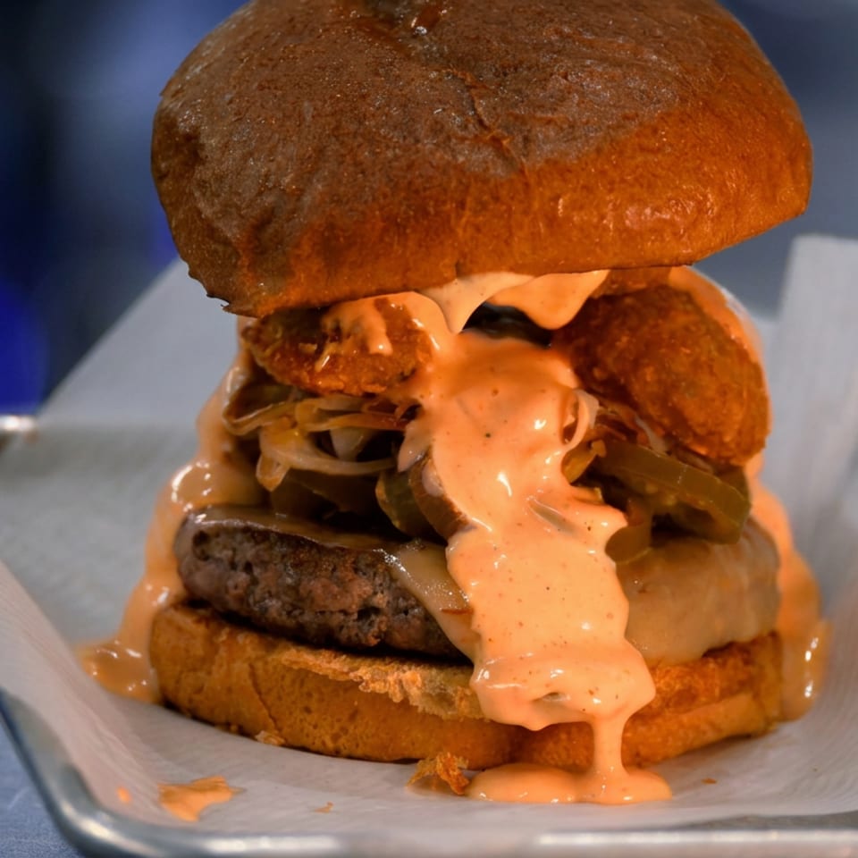 Close-up of a loaded double burger with fried toppings, melted cheese, onions, and creamy sauce on parchment paper.