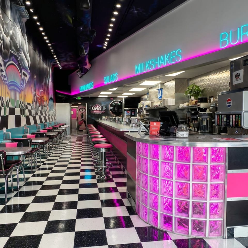 Retro diner interior with black-and-white checkered floors, turquoise booths, pink stools, neon signage, and a chrome service counter.