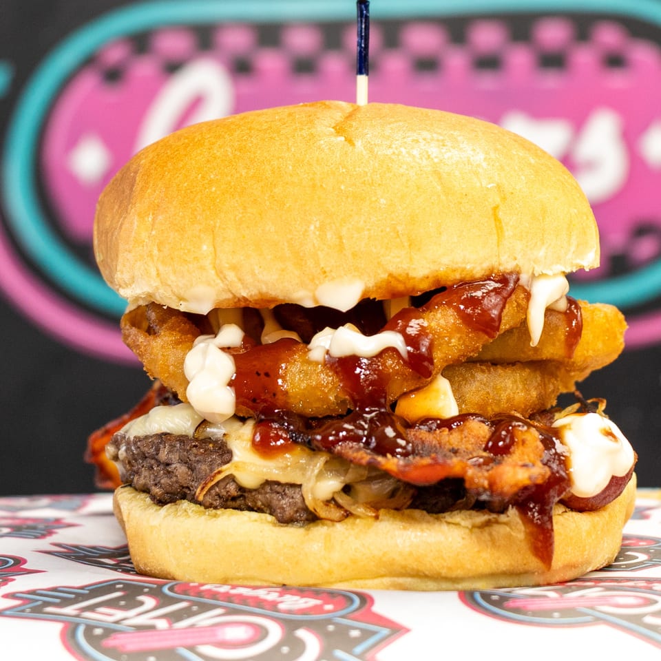 Towering bacon cheeseburger with fried toppings, barbecue sauce, mayonnaise, and a skewer, displayed against a colorful diner-style backdrop.