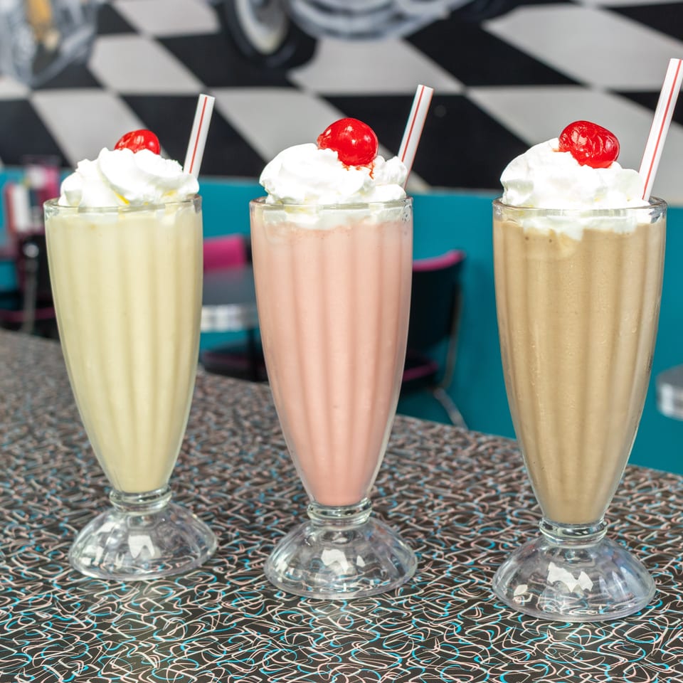 Three colorful milkshakes topped with whipped cream, cherries, and striped straws on a retro diner table.
