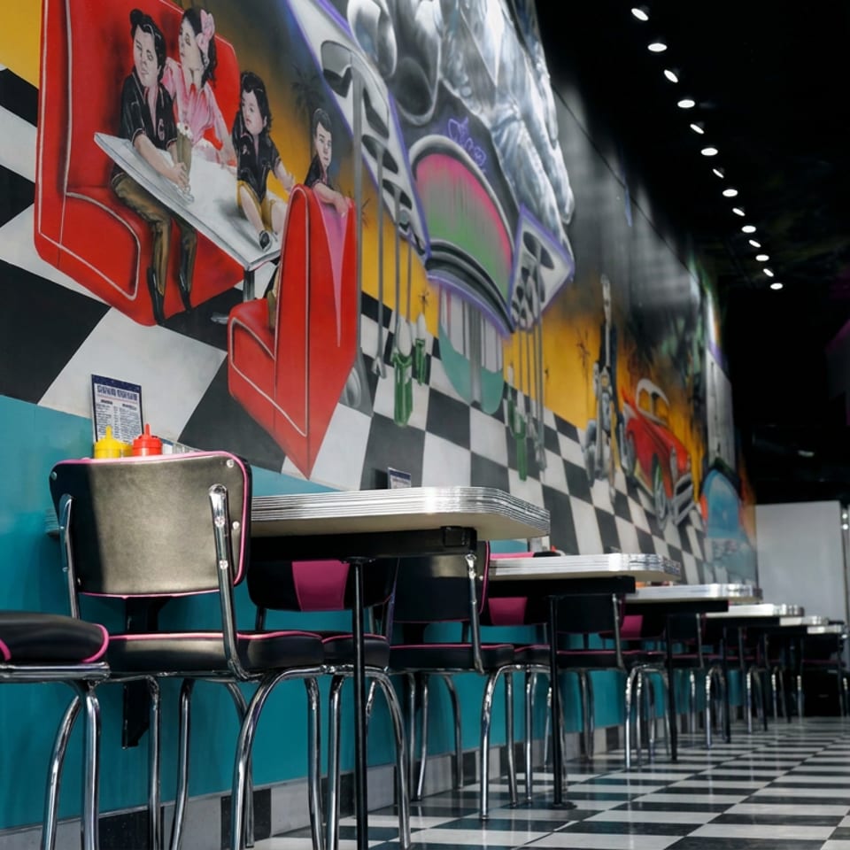 Retro diner interior with chrome-edged booths, checkerboard floor, colorful murals, and rows of tables and chairs.
