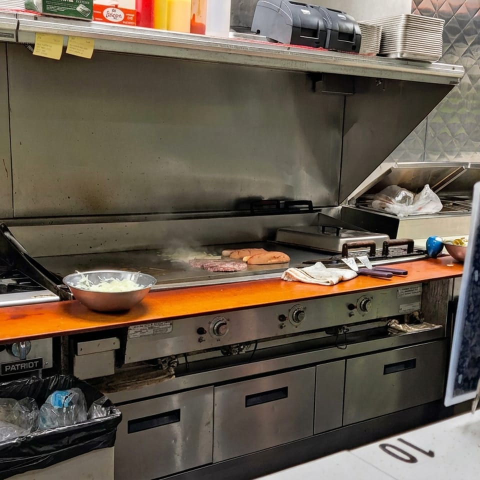 Commercial restaurant kitchen with a stainless-steel grill, cooking meat, a bowl of chopped onions, utensils, and stacked plates.